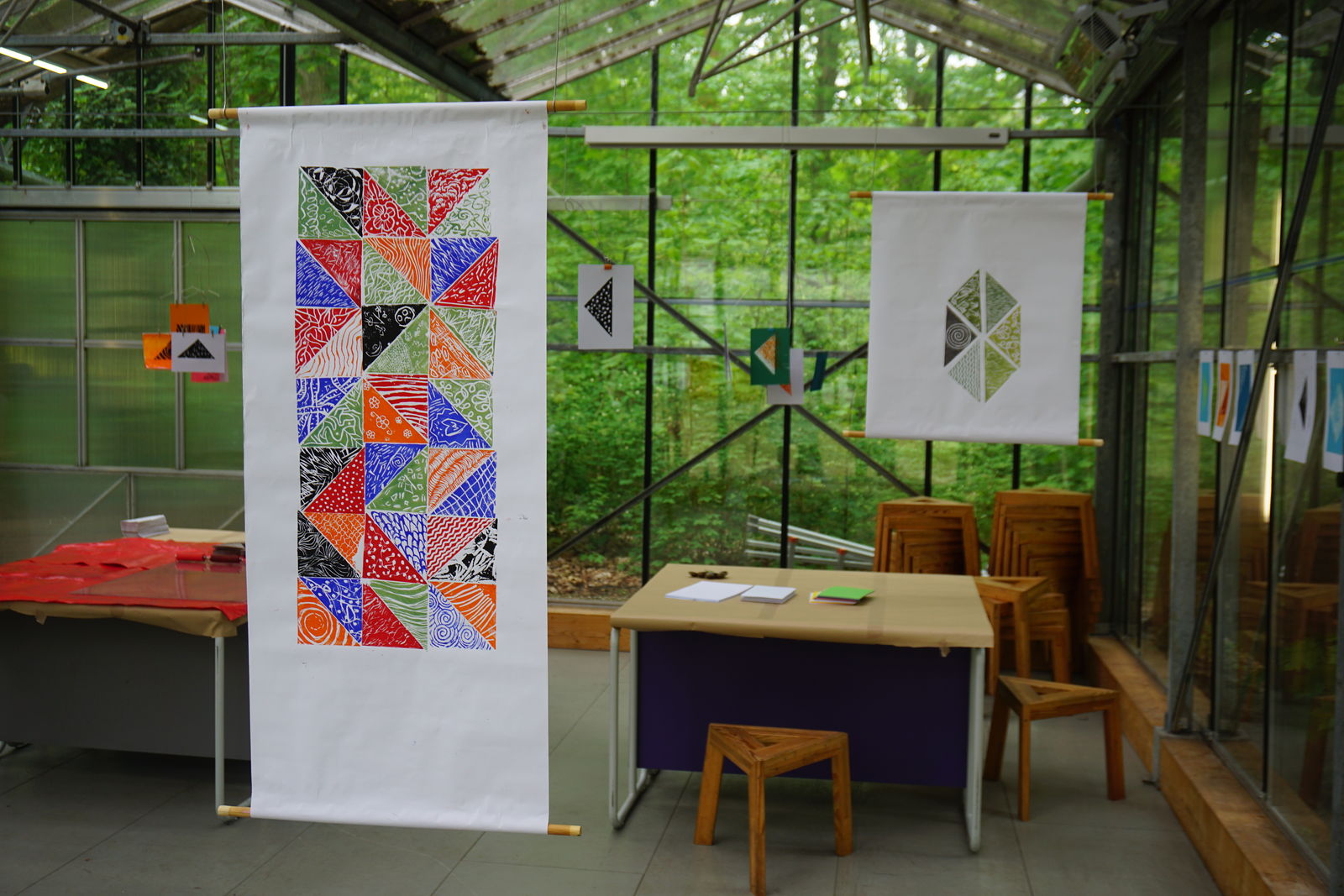 Foto, Eine farbenfrohe Textilkunst-Ausstellung mit handgemusterten Stoffbahnen und geometrischen Motiven ist in einem lichtdurchfluteten Gewächshaus zu sehen